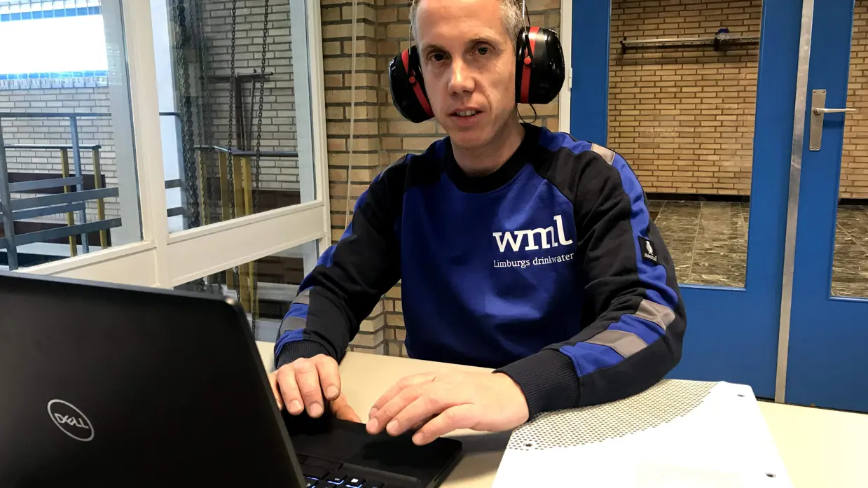 Jeroen Blomen met gehoorbescherming achter zijn laptop met een trui van Limburgse Waterleidingen aan. 