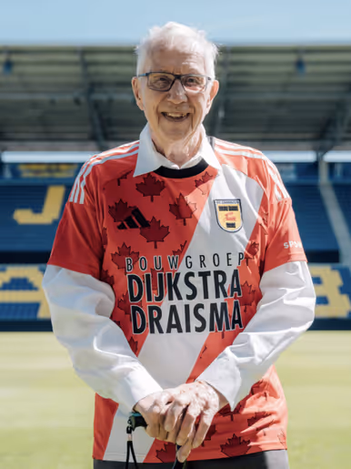 Een glimlachende oudere man staat op een voetbalveld, gekleed in een rood-wit shirt met esdoornbladeren en de tekst "Dijkstra Draisma" en "Bouwgroep" erop.