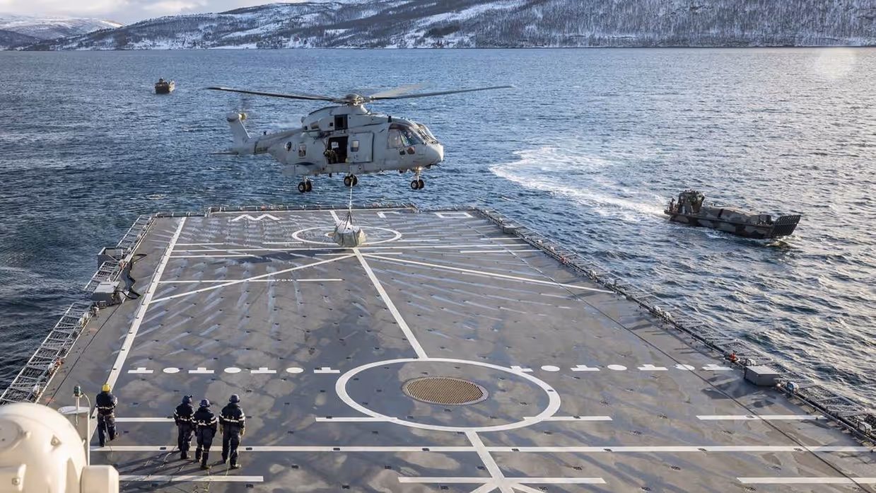 Helikopter tilt vracht boven landingsdek marineschip. Landingsvaartuig vaart langszij.