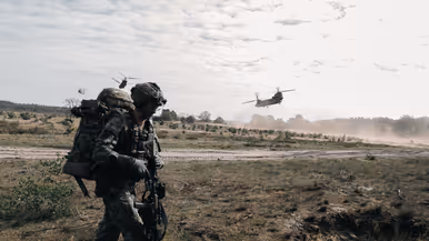 Een militair met zware bepakking verplaatst zich over de heide terwijl Chinook-helikopters laag overvliegen.