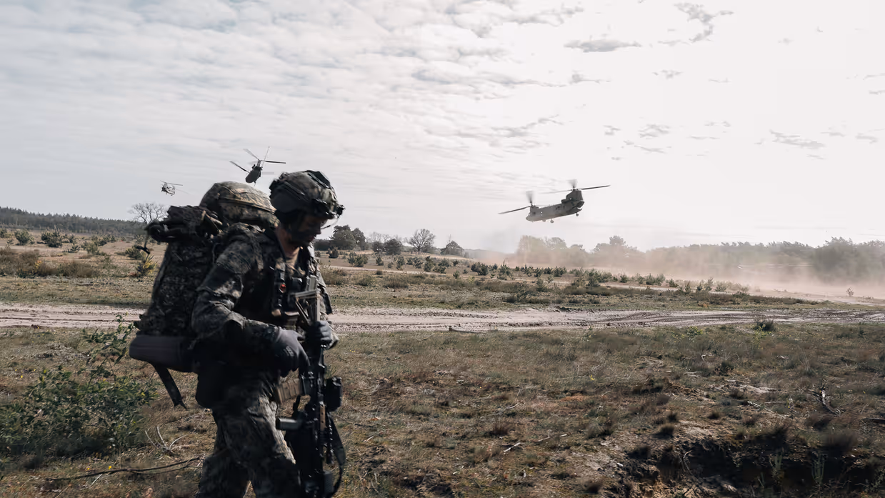 Een militair met zware bepakking verplaatst zich over de heide terwijl Chinook-helikopters laag overvliegen.