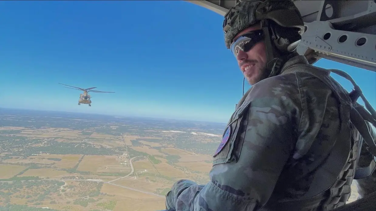 Naar de camera omkijkende militair met zonnebril en helm op zittend op laadklep van vliegende Chinook. Op de achtergrond vliegt ook een Chinook.