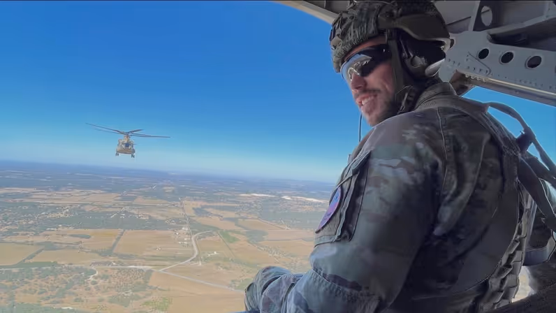Naar de camera omkijkende militair met zonnebril en helm op zittend op laadklep van vliegende Chinook. Op de achtergrond vliegt ook een Chinook.