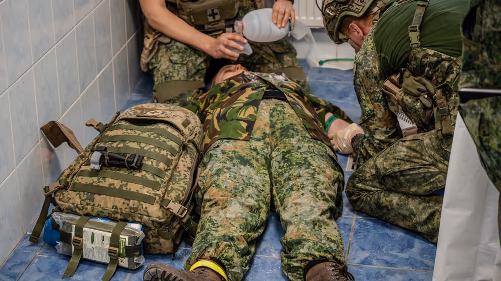 Gewonde militair ligt op zijn rug op de grond. Hij wordt behandeld.