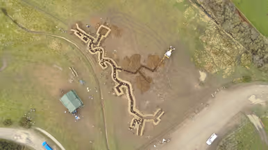 Links: Luchtfoto van loopgraven op militair oefenterrein. Rechts: Militair met wapen hurkt in loopgraaf waarvan de wanden met golfplaten zijn bekleed. Op zijn helm een bos stro als camouflage.