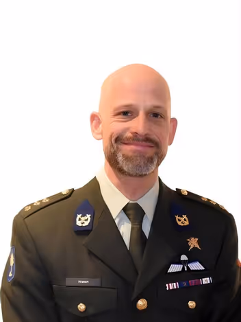 Een glimlachende, kaalgeschoren man in militair uniform met onderscheidingen, insignes, en een das, draagt op zijn borst een naamkaartje met ‘Nissen’. 