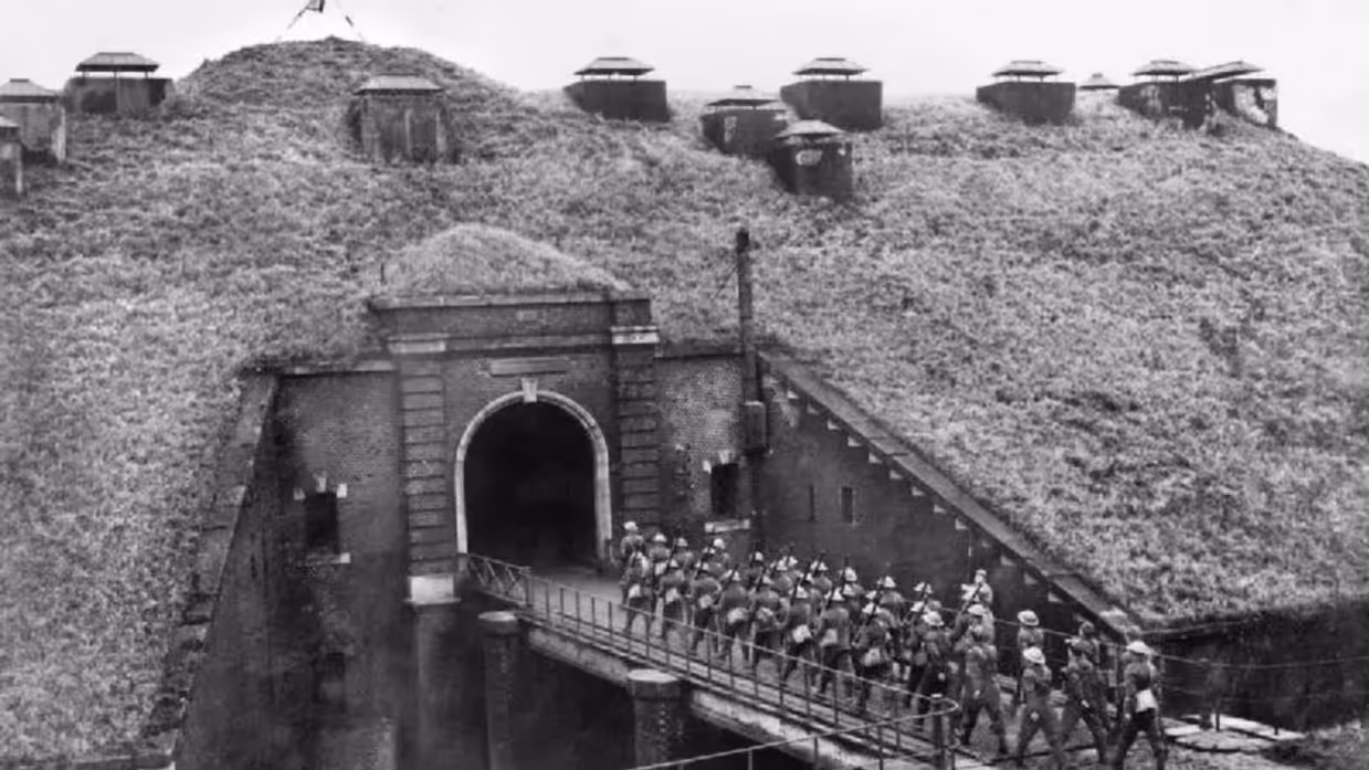 Een groep soldaten loopt over een smalle brug naar een betonnen tunnel in een groene heuvel met bunkers.