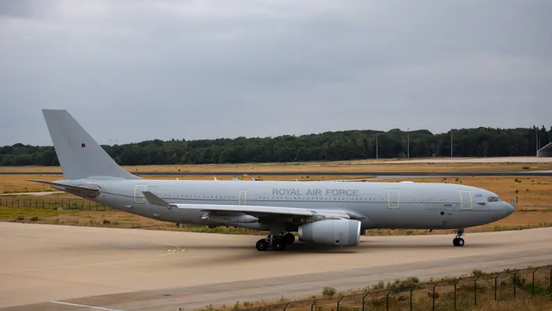 Een grijs militair vliegtuig met "Royal Air Force" op de romp staat op een startbaan, omringd door gras en bomen.