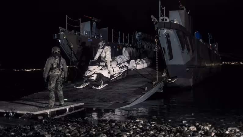  Een marinier in wit pak bestuurt een sneeuwscooter. Hiermee verlaat hij, in het donker, een landingsvaartuig.