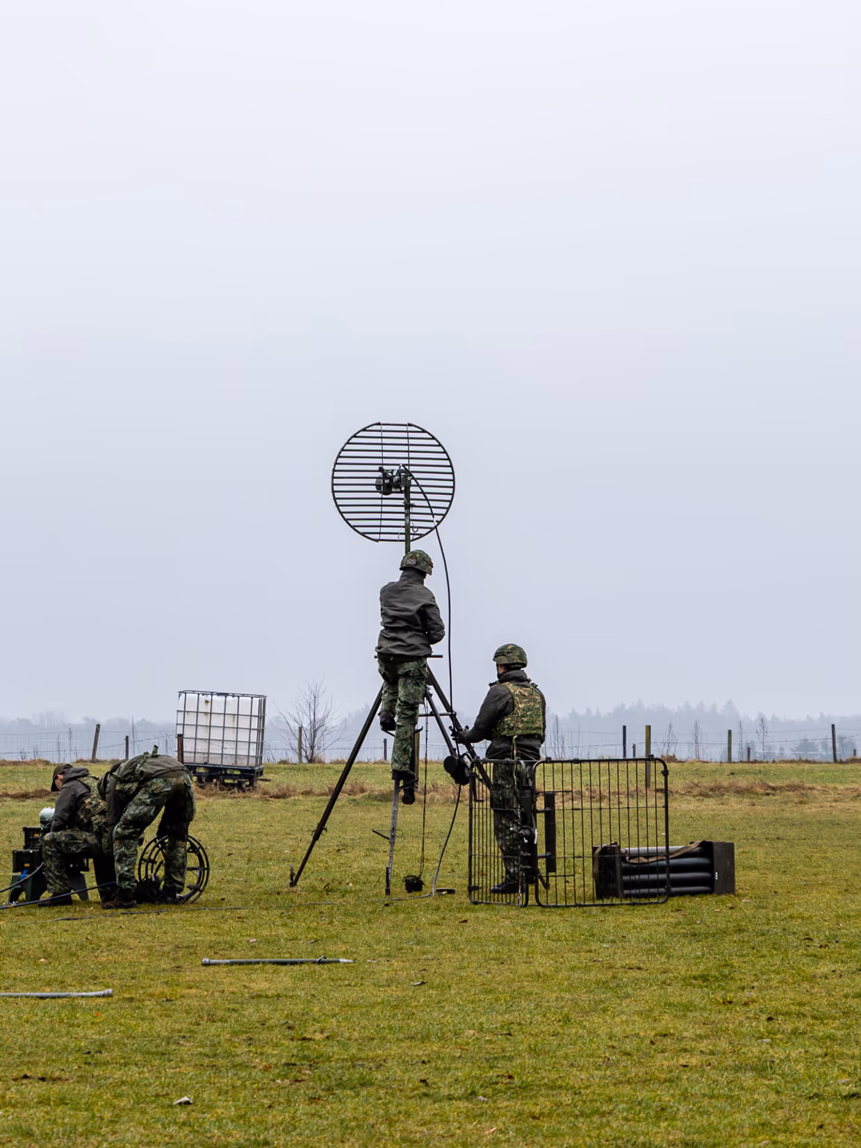  foto links: Militairen installeren een ronde satellietschotel, omringd door uitrusting en voertuigen. Links midden: Een groep soldaten loopt over een keienpad langs een station met een overdekt terras; een soldaat loopt voorop, de rest volgt in een rij. Rechts midden: Drie militairen werken op een open veld: één stelt een grote satellietschotel af, twee anderen bedienen apparatuur en kabels op de grond. Rechts: Vier militairen in gevechtstenue lopen over een grasveld langs een onverharde weg;