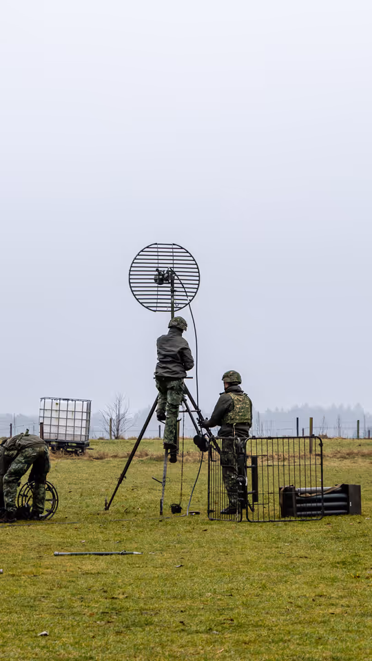  foto links: Militairen installeren een ronde satellietschotel, omringd door uitrusting en voertuigen. Links midden: Een groep soldaten loopt over een keienpad langs een station met een overdekt terras; een soldaat loopt voorop, de rest volgt in een rij. Rechts midden: Drie militairen werken op een open veld: één stelt een grote satellietschotel af, twee anderen bedienen apparatuur en kabels op de grond. Rechts: Vier militairen in gevechtstenue lopen over een grasveld langs een onverharde weg;