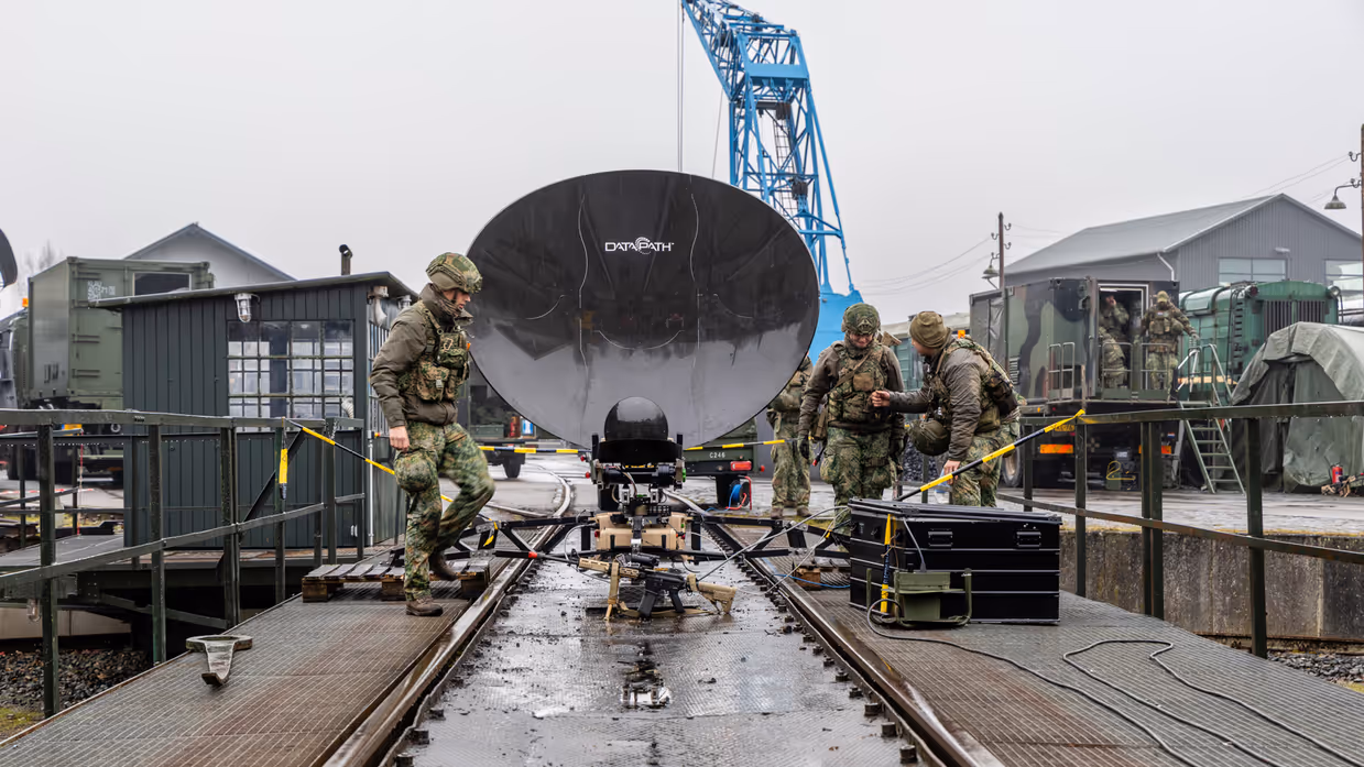  foto links: Militairen installeren een ronde satellietschotel, omringd door uitrusting en voertuigen. Links midden: Een groep soldaten loopt over een keienpad langs een station met een overdekt terras; een soldaat loopt voorop, de rest volgt in een rij. Rechts midden: Drie militairen werken op een open veld: één stelt een grote satellietschotel af, twee anderen bedienen apparatuur en kabels op de grond. Rechts: Vier militairen in gevechtstenue lopen over een grasveld langs een onverharde weg;