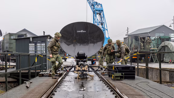  foto links: Militairen installeren een ronde satellietschotel, omringd door uitrusting en voertuigen. Links midden: Een groep soldaten loopt over een keienpad langs een station met een overdekt terras; een soldaat loopt voorop, de rest volgt in een rij. Rechts midden: Drie militairen werken op een open veld: één stelt een grote satellietschotel af, twee anderen bedienen apparatuur en kabels op de grond. Rechts: Vier militairen in gevechtstenue lopen over een grasveld langs een onverharde weg;