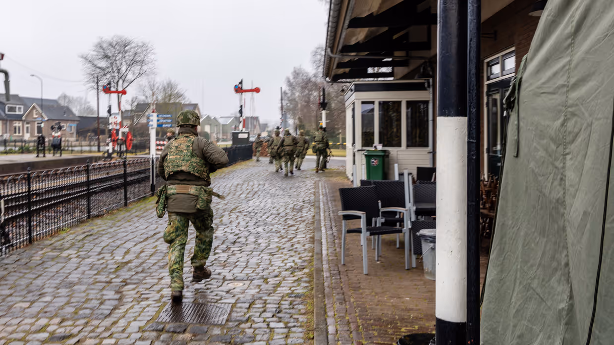  foto links: Militairen installeren een ronde satellietschotel, omringd door uitrusting en voertuigen. Links midden: Een groep soldaten loopt over een keienpad langs een station met een overdekt terras; een soldaat loopt voorop, de rest volgt in een rij. Rechts midden: Drie militairen werken op een open veld: één stelt een grote satellietschotel af, twee anderen bedienen apparatuur en kabels op de grond. Rechts: Vier militairen in gevechtstenue lopen over een grasveld langs een onverharde weg;