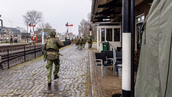  foto links: Militairen installeren een ronde satellietschotel, omringd door uitrusting en voertuigen. Links midden: Een groep soldaten loopt over een keienpad langs een station met een overdekt terras; een soldaat loopt voorop, de rest volgt in een rij. Rechts midden: Drie militairen werken op een open veld: één stelt een grote satellietschotel af, twee anderen bedienen apparatuur en kabels op de grond. Rechts: Vier militairen in gevechtstenue lopen over een grasveld langs een onverharde weg;