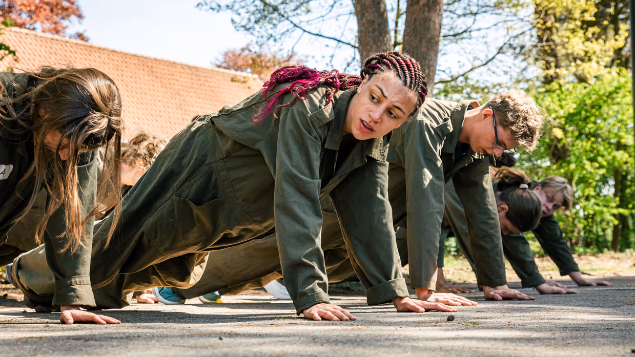 Een groep MDT-deelnemers doet samen een fysieke oefening (push-ups) op de kazerne, waarbij ze elkaar motiveren en samenwerken. 