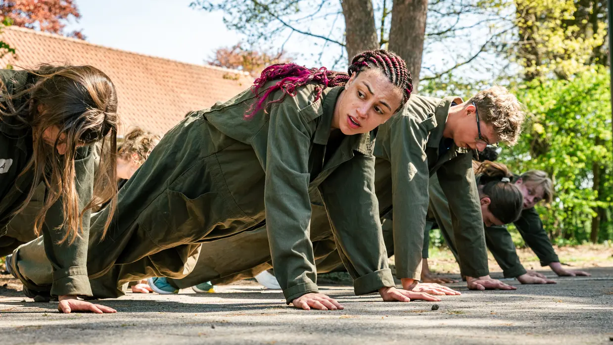 Een groep MDT-deelnemers doet samen een fysieke oefening (push-ups) op de kazerne, waarbij ze elkaar motiveren en samenwerken. 