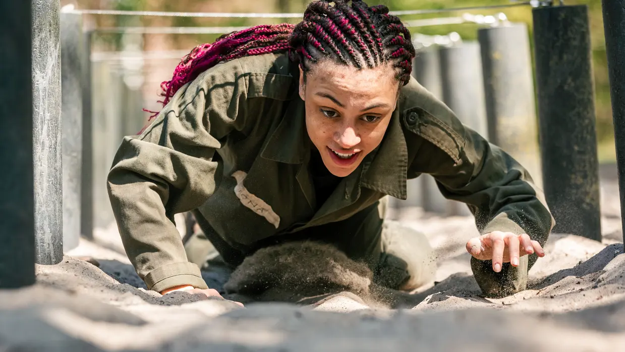 MDT-deelnemer Fiona kruipt onder lage hindernissen door in het zand tijdens een militaire oefening op de kazerne. 