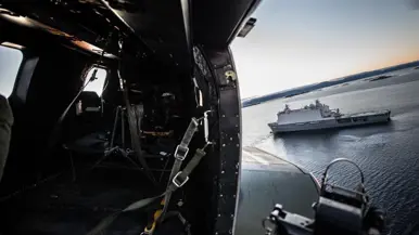 Zicht op een marineschip vanuit de open deur van een voorbijvliegende helikopter.