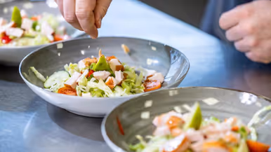 Op de linkerfoto voegen twee handen ingrediënten toe aan salades. Op de rechterfoto kijkt generaal Jobse toe hoe één van de cursisten een stuk groente snijdt. 