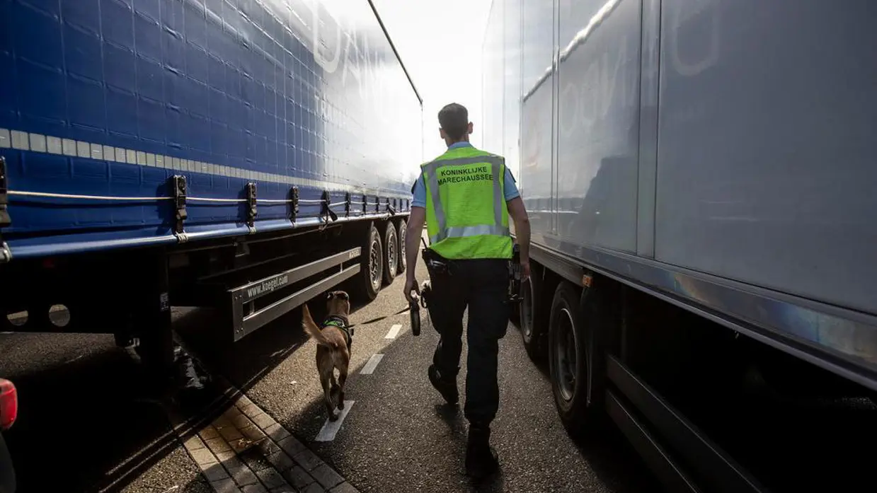 Een marechaussee loopt met zijn speurhond tussen twee vrachtwagens. 