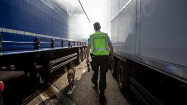 Een marechaussee loopt met zijn speurhond tussen twee vrachtwagens. 
