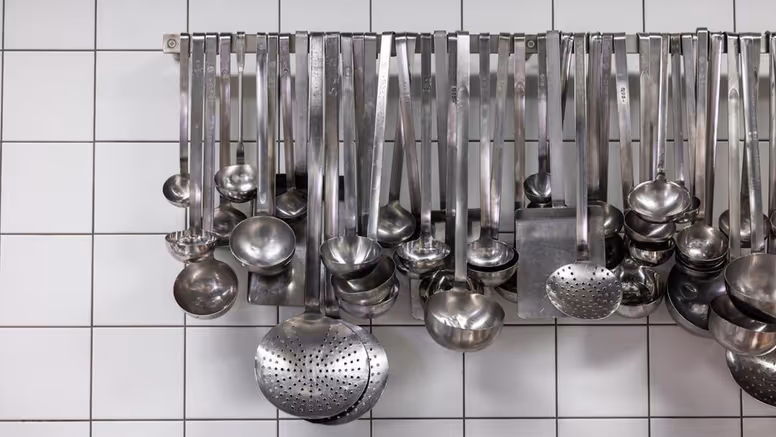 Grote lepels en ander staalkleurig keukengerei hangt aan een rek op een witte tegelmuur. 