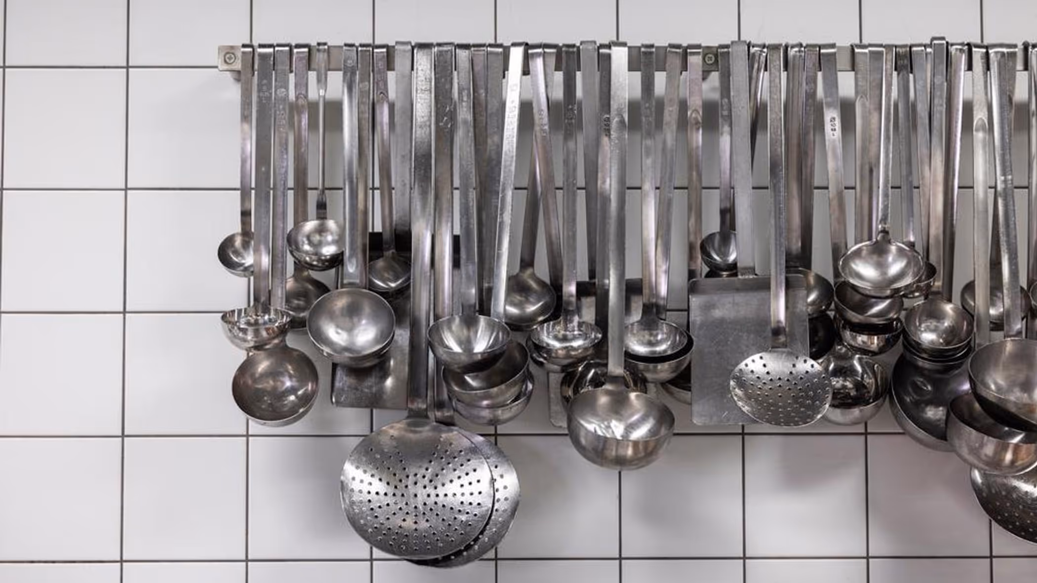 Grote lepels en ander staalkleurig keukengerei hangt aan een rek op een witte tegelmuur. 