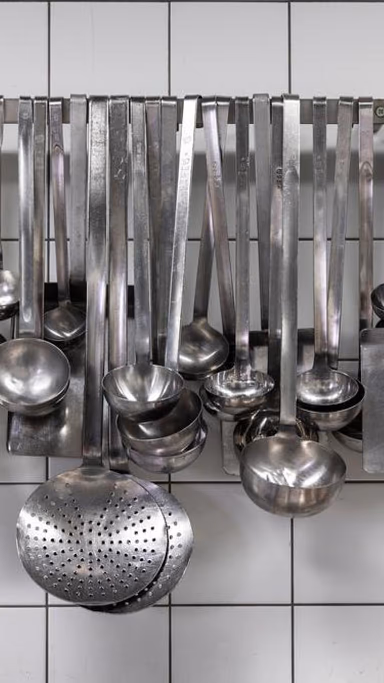 Grote lepels en ander staalkleurig keukengerei hangt aan een rek op een witte tegelmuur. 