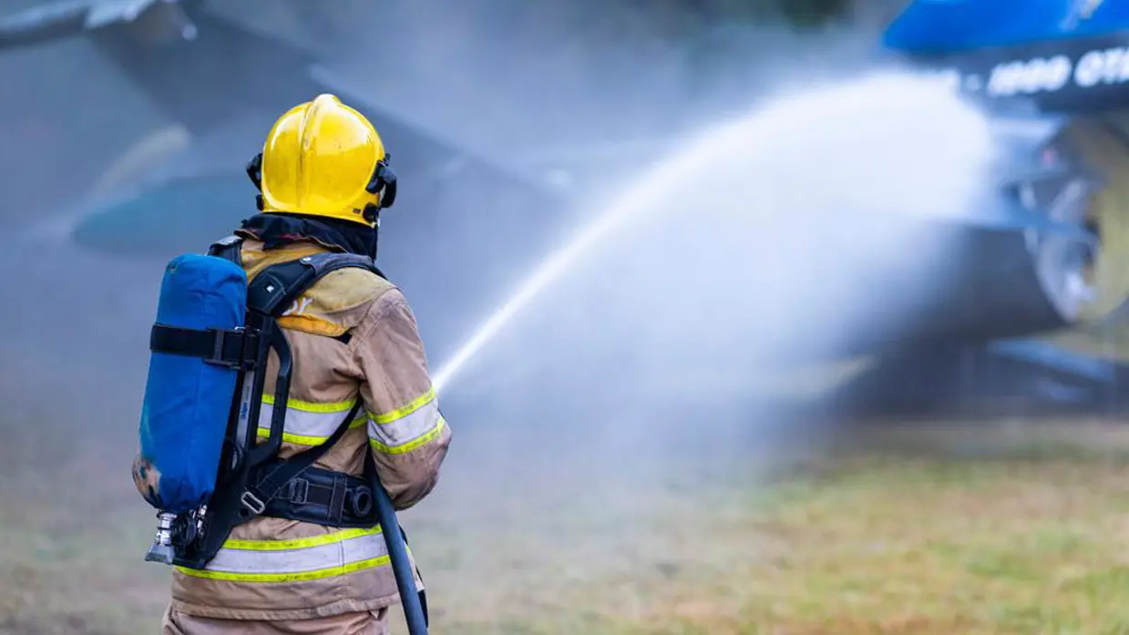 Een brandweerman met een fles perslucht op zijn rug richt een waterstraal op het wrak van een jachtvliegtuig.
