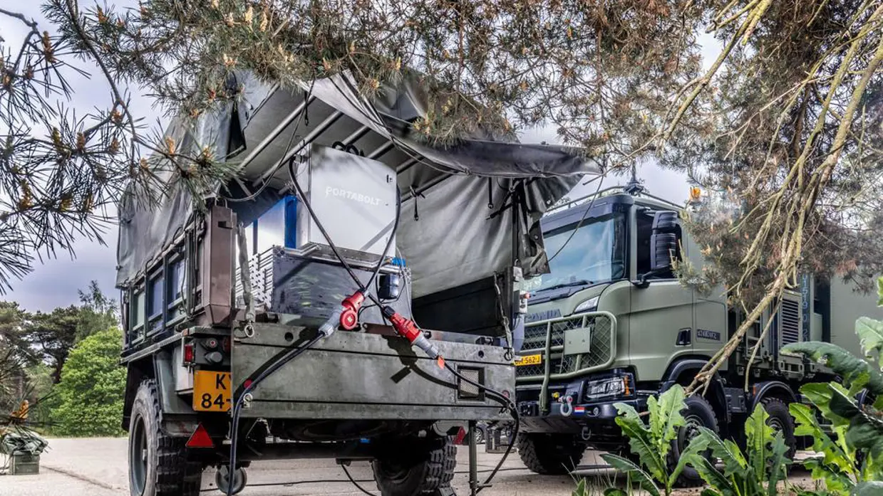 Een aanhangwagen in camouflagekleuren staat in het groen. In de aanhangwagen staat een zilveren batterijsysteem.