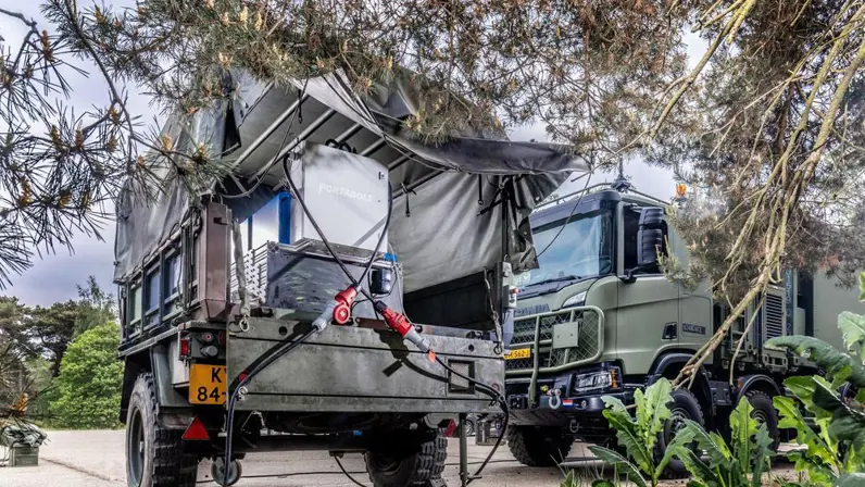 Een aanhangwagen in camouflagekleuren staat in het groen. In de aanhangwagen staat een zilveren batterijsysteem.