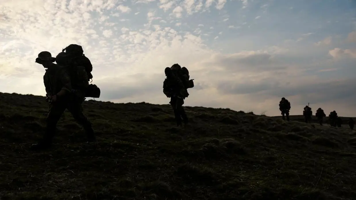 Silhouetten van mariniers met zware bepakking lopend door een heuvellandschap.