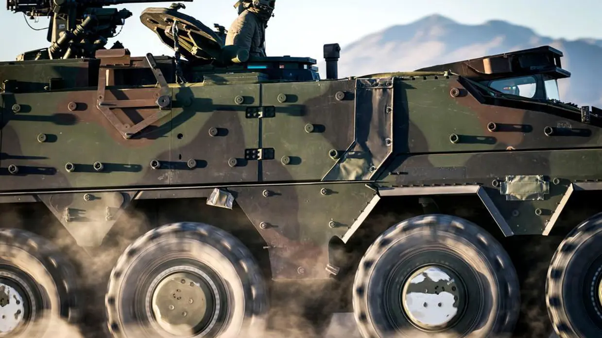Een Boxer-pantservoertuigen op wielen raast over een zandvlakte. Bovenop zit een militair in een openstaand luik.