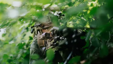 Een militair ligt achter zijn geweer in het bos.