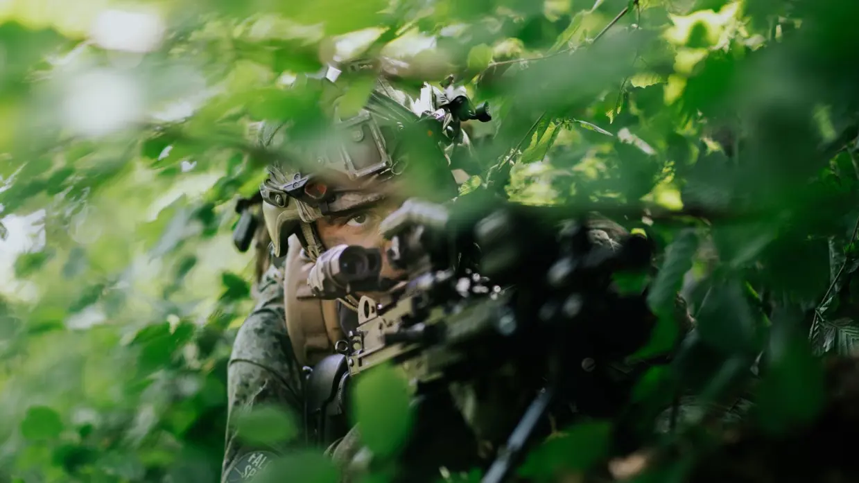 Een militair ligt achter zijn geweer in het bos.