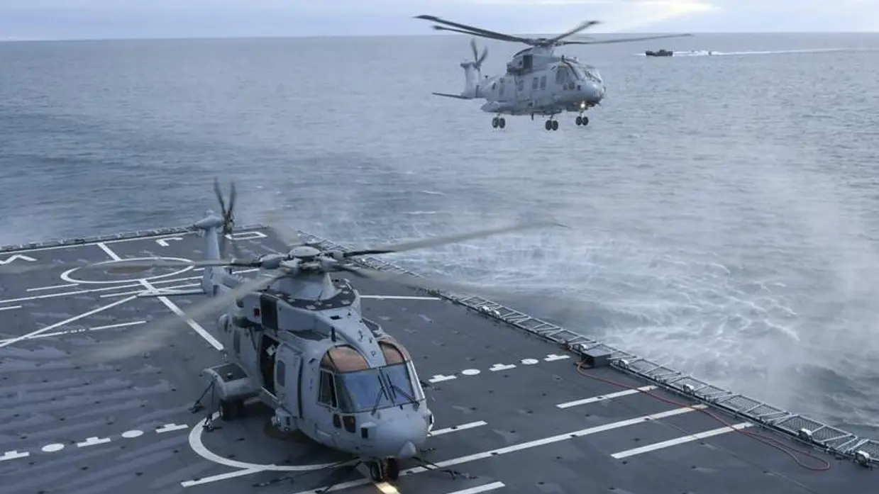 Een helikopter vliegt boven landingsdek schip. Een ander toestel staat op het dek.