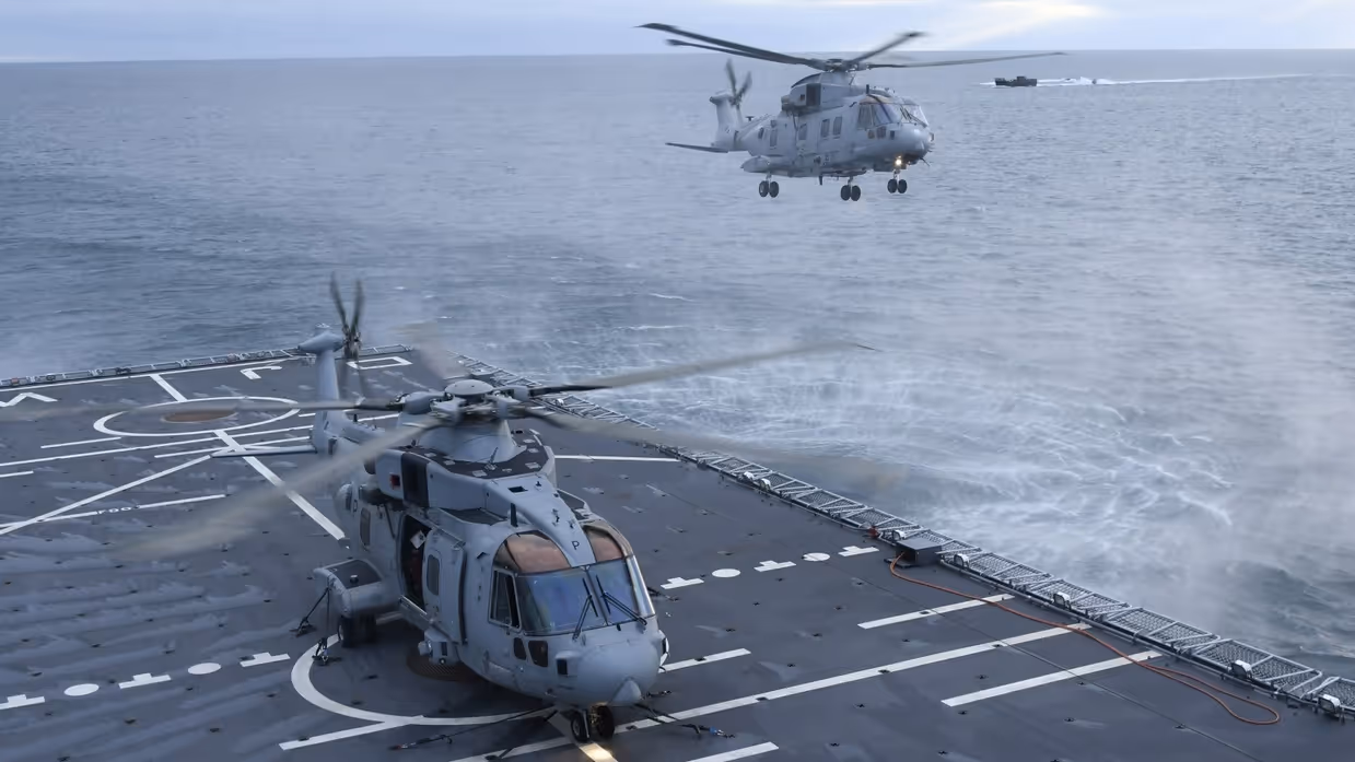 Een grijze helikopter is zojuist geland op het helikopterdek van een marineschip. Daarachter hangt een tweede toestel net boven het water.