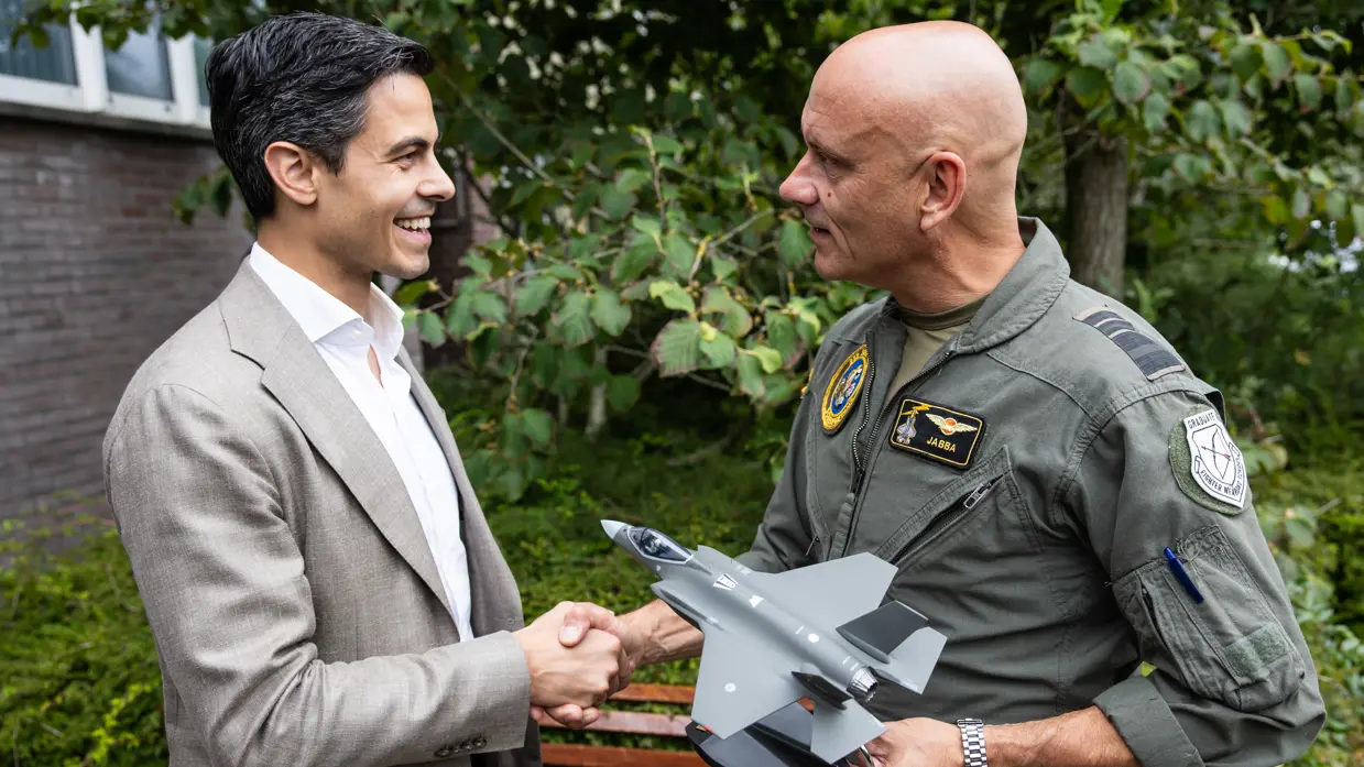 Steur schudt Jetten de hand terwijl hij hem een miniatuur van een F-35 overhandigd. Op de achtergrond bossage en een houten bankje naast een stenen gebouw.
