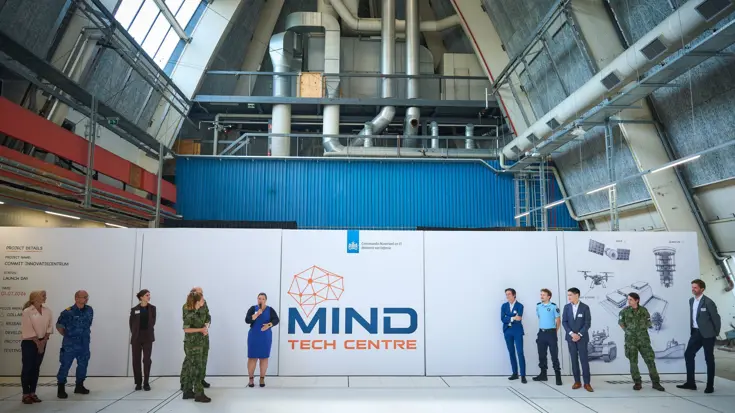 Een witte muur met daarop de tekst ‘MIND Tech Centre’ in een hoge, grijze ruimte. Aan weerszijden van deze tekst staan militairen en burgers, van wie één met microfoon in de hand.
