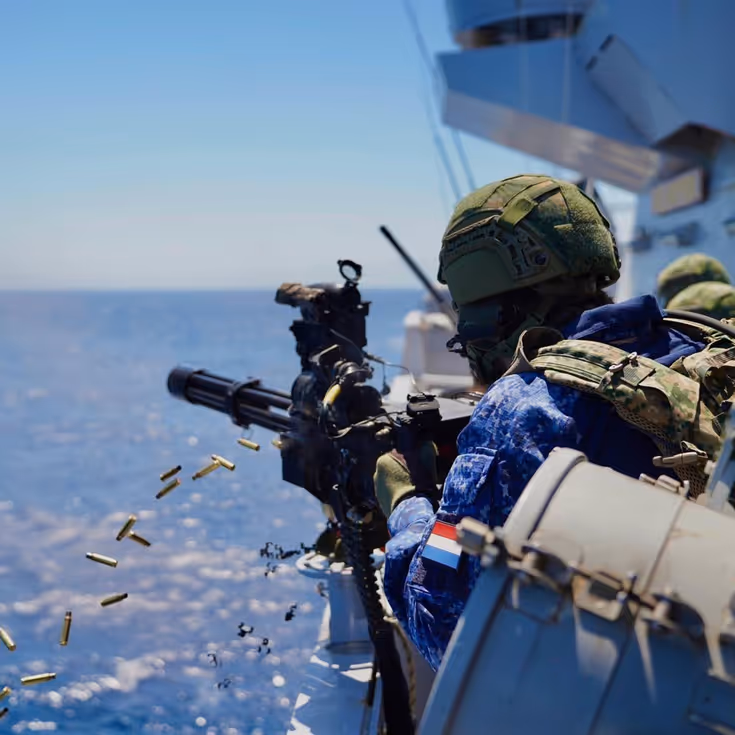 Een militair met helm op en scherfvest aan vuurt met de minigun die is gemonteerd op de reling van het schip. Hulzen vliegen in het rond.