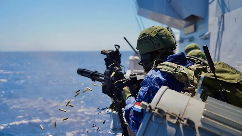 Een marineman vuurt met een minigun vanaf de reling van een dek.