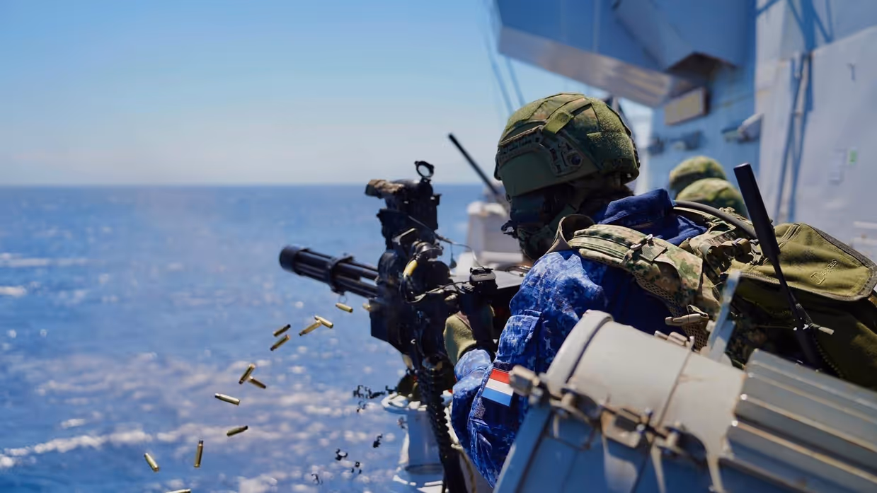 Een militair met helm op en scherfvest aan vuurt met de minigun die is gemonteerd op de reling van het schip. Hulzen vliegen in het rond.