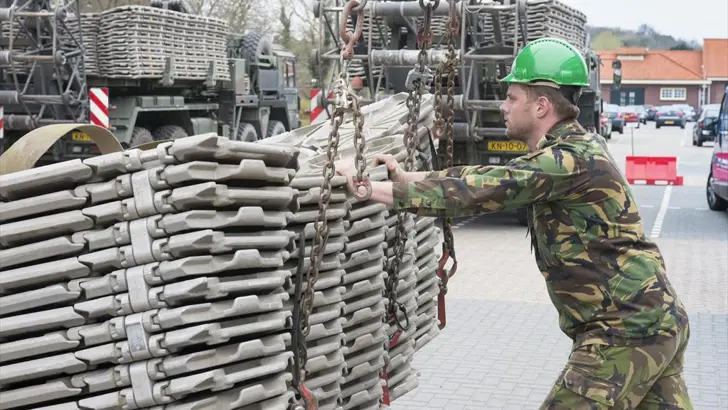 Een militair duwt tegen een stapel wegenmatten. De stapel hangt aan een hijskraan.