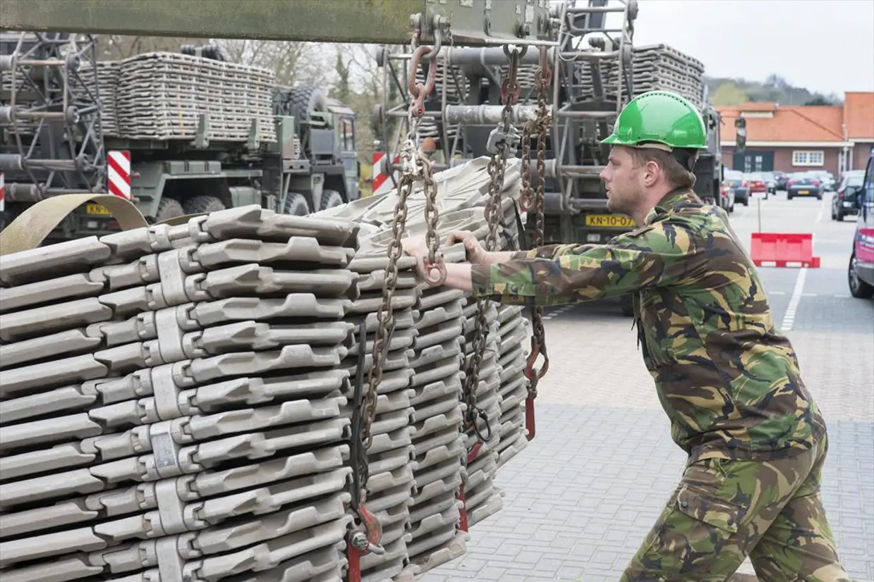 Een militair duwt tegen een stapel wegenmatten. De stapel hangt aan een hijskraan.