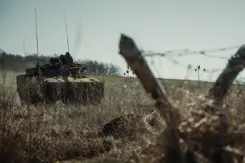 Een tank op een veld. Op de voorgrond 2 schuine palen in V-vorm met prikkeldraad.