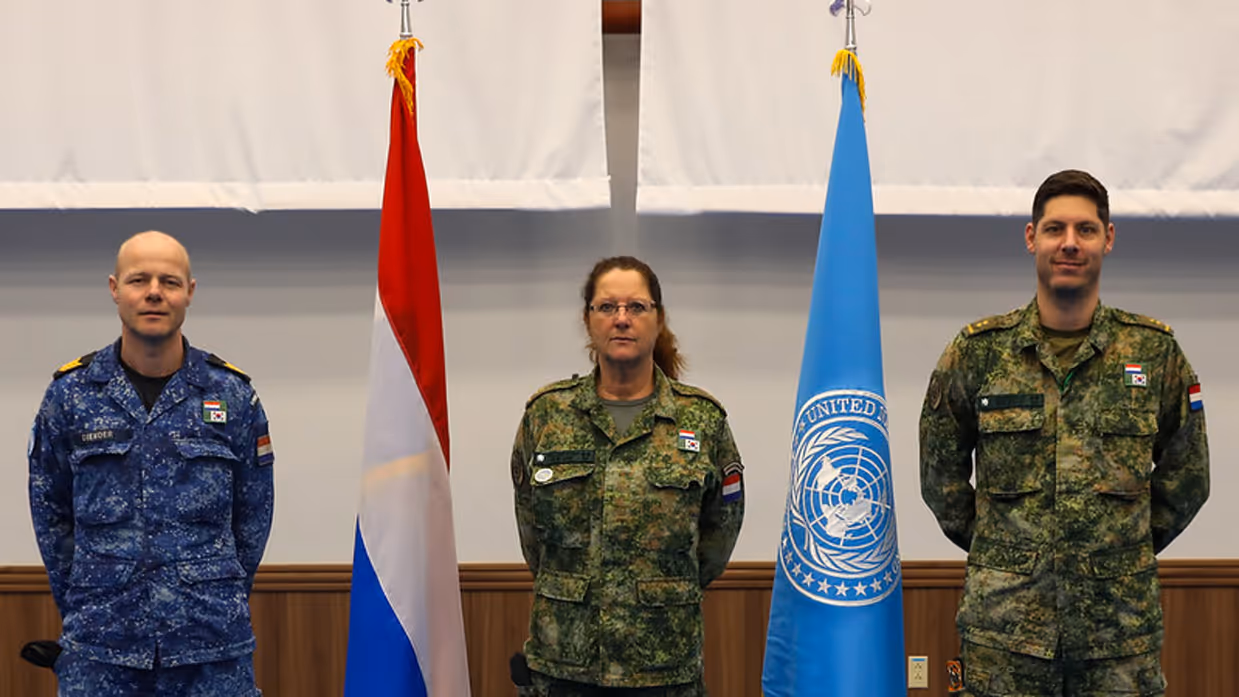 3 militairen staan opgesteld naast een Nederlandse vlag en naast een vlag van de NAVO. Twee dragen een groen gevechtspak, Leon Diender het blauwe boordtenue.&nbsp;