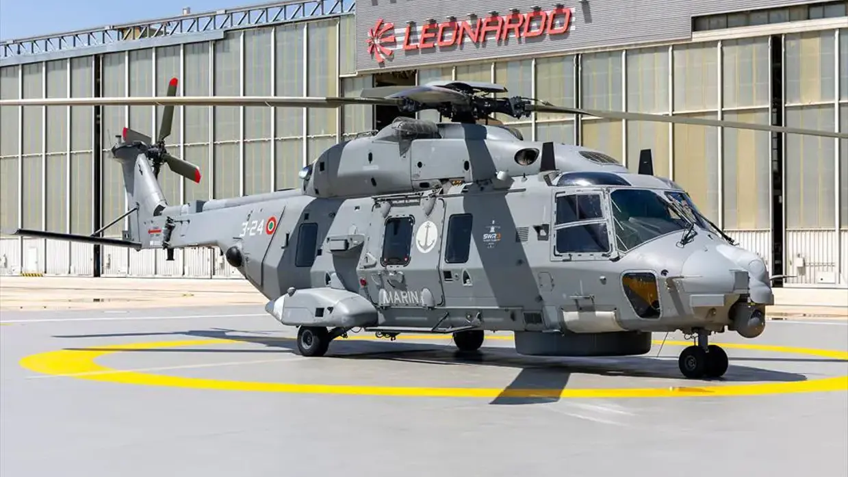 Een grijze NH90-helikopter staat op een platform. Daarachter een hangaar mat de tekst ‘Leonardo’. 