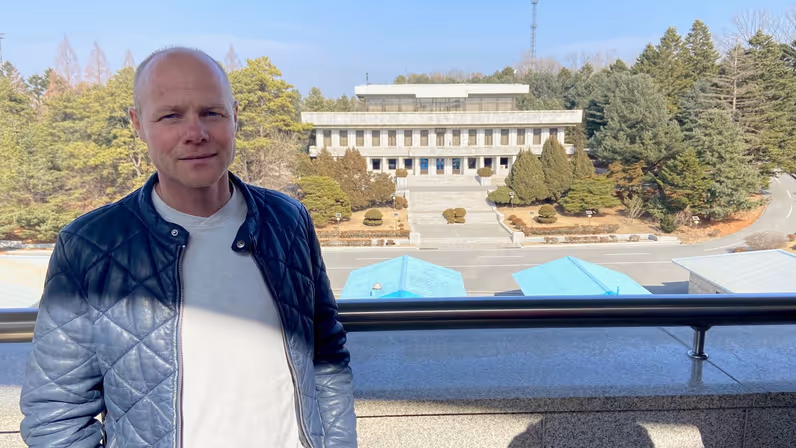 Leon Diender in een blauwe jas op een balkon dat uitkijkt over blauwe gebouwtjes op de grens van de gedemilitariseerde zone.&nbsp;