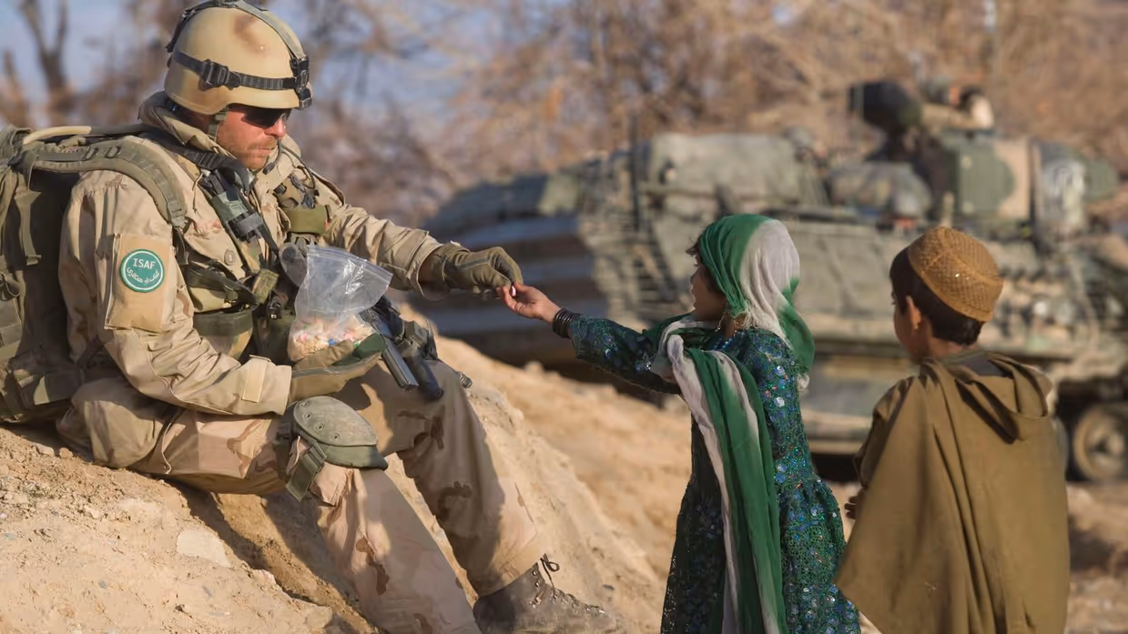 Alt-tekst: Een militair zit op een verhoging van zand en geeft een snoepje aan een meisje. Achter haar staat een jongen. 